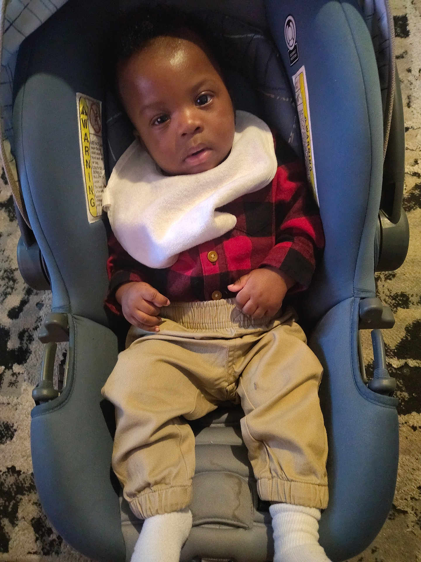 Kingston S Hicks is registered to the contest to win money with this photo: baby, infant, child, toddler, car_seat, bib, plaid_shirt, checkered_shirt, khaki_pants, socks, hands, face, eyes, looking_at_camera, indoor, carpet, floor, cozy, portrait, dark_hair