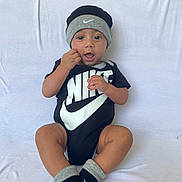 Gustavo is registered to the contest to win money with this photo: baby, child, infant, black_clothing, nike_logo, hat, socks, onesie, sitting, white_background, cute, smiling, hand_near_mouth, indoors, portrait, young_child, fashion, footwear, headwear, expression