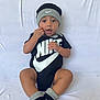baby, child, infant, black_clothing, nike_logo, hat, socks, onesie, sitting, white_background, cute, smiling, hand_near_mouth, indoors, portrait, young_child, fashion, footwear, headwear, expression