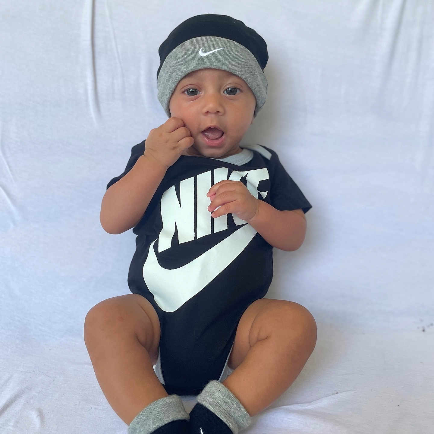 Gustavo is registered to the contest to win money with this photo: baby, black_clothing, child, cute, expression, fashion, footwear, hand_near_mouth, hat, headwear, indoors, infant, nike_logo, onesie, portrait, sitting, smiling, socks, white_background, young_child