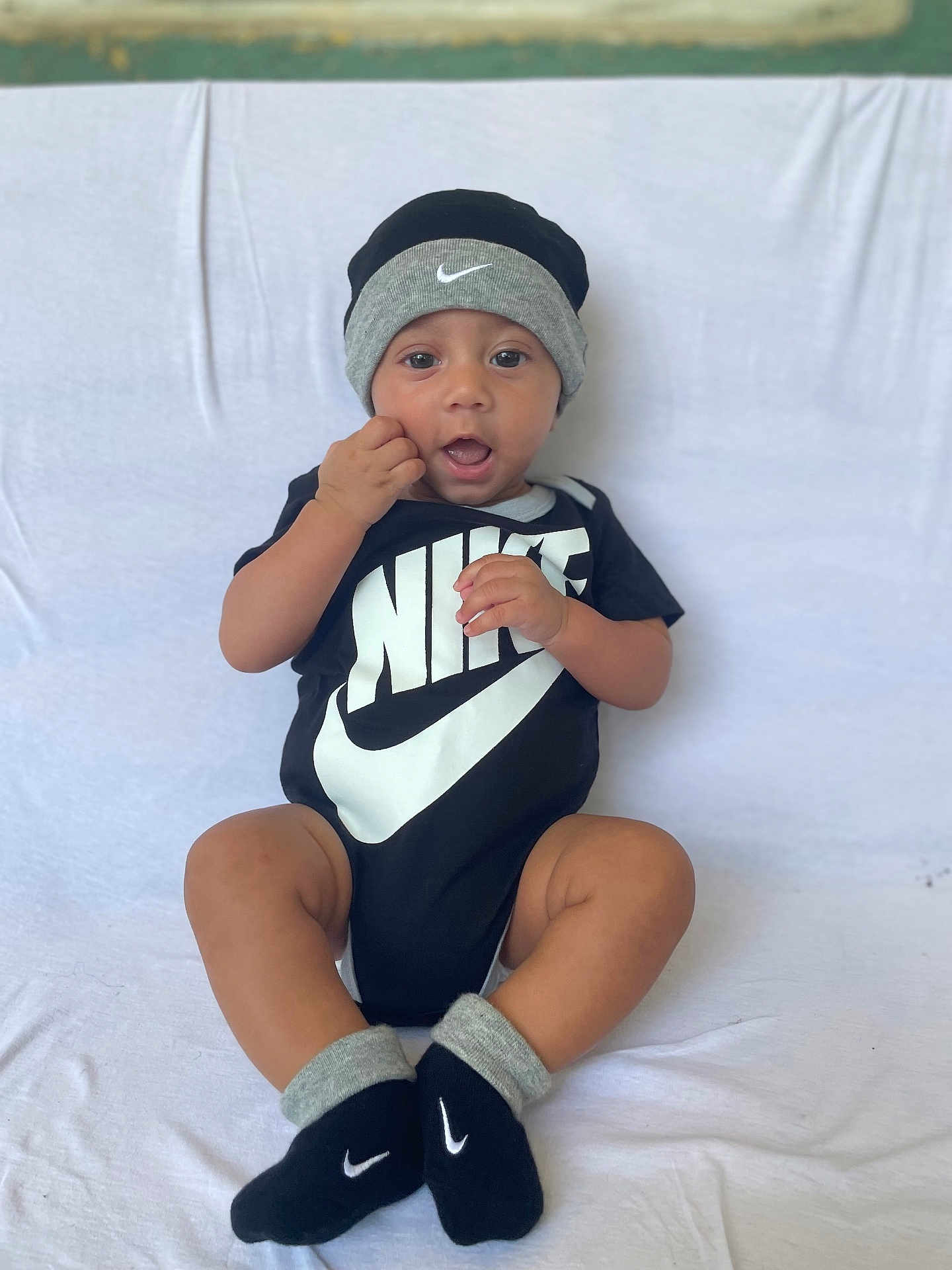 Gustavo is registered to the contest to win money with this photo: baby, child, infant, black_clothing, nike_logo, hat, socks, onesie, sitting, white_background, cute, smiling, hand_near_mouth, indoors, portrait, young_child, fashion, footwear, headwear, expression