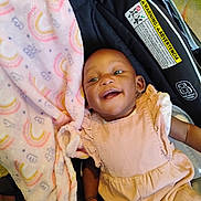 Evvie is registered to the contest to win money with this photo: baby, smiling, infant, car_seat, blanket, pink_blanket, onesie, ruffle_dress, happy, face, hand, safety_label, warning_label, strap, stroller, shoe, floor, timestamp, watermark, portrait