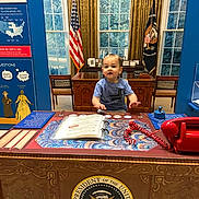 Tyler is registered to the contest to win money with this photo: toddler, child, desk, presidential_seal, flag, american_flag, phone, book, curtains, window, chair, indoor, museum, historical_display, red_telephone, blue_wall, wooden_furniture, patriotic, official, playful