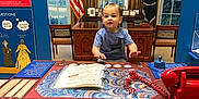 Tyler is registered to the contest to win money with this photo: american_flag, blue_wall, book, chair, child, curtains, desk, flag, historical_display, indoor, museum, official, patriotic, phone, playful, presidential_seal, red_telephone, toddler, window, wooden_furniture