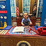 toddler, child, desk, presidential_seal, flag, american_flag, phone, book, curtains, window, chair, indoor, museum, historical_display, red_telephone, blue_wall, wooden_furniture, patriotic, official, playful