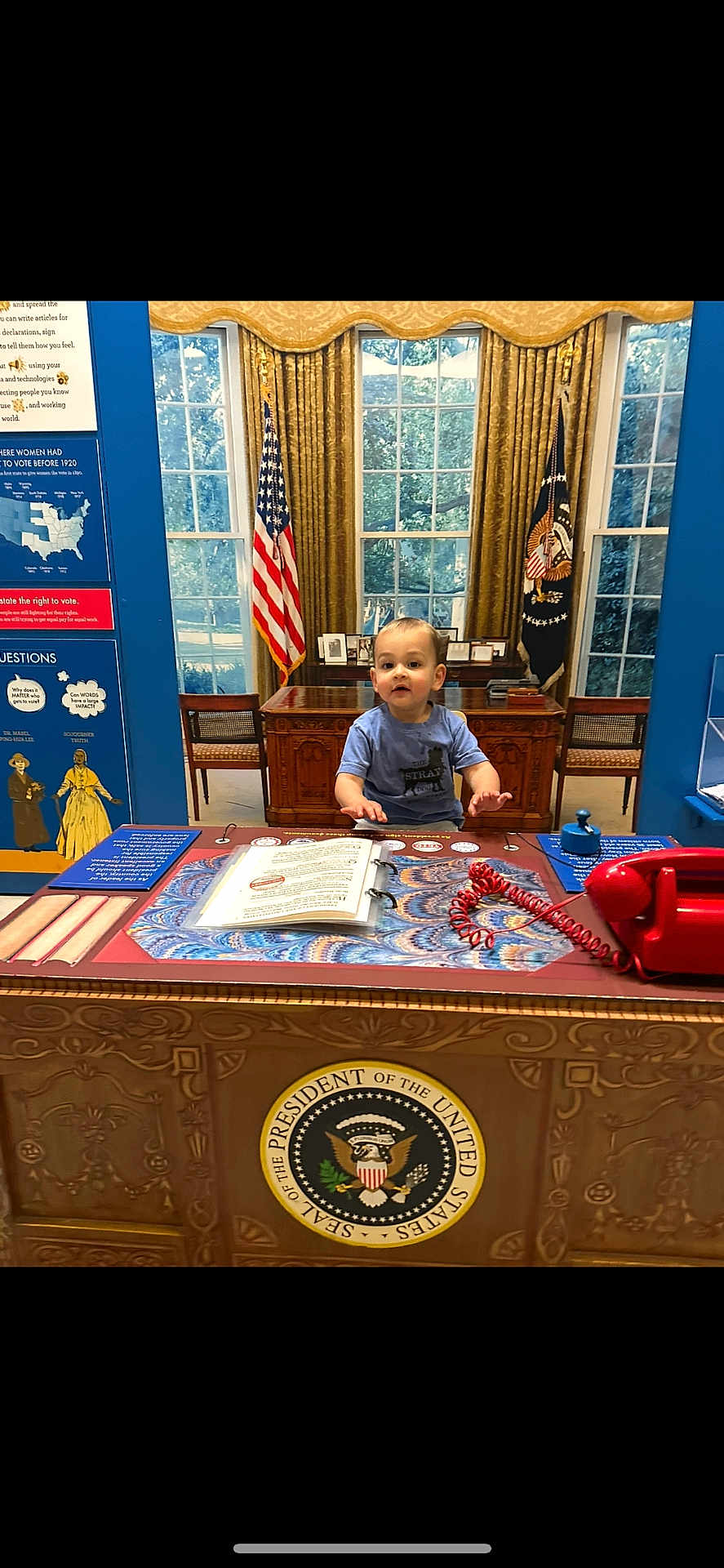 Tyler is registered to the contest to win money with this photo: toddler, child, desk, presidential_seal, flag, american_flag, phone, book, curtains, window, chair, indoor, museum, historical_display, red_telephone, blue_wall, wooden_furniture, patriotic, official, playful