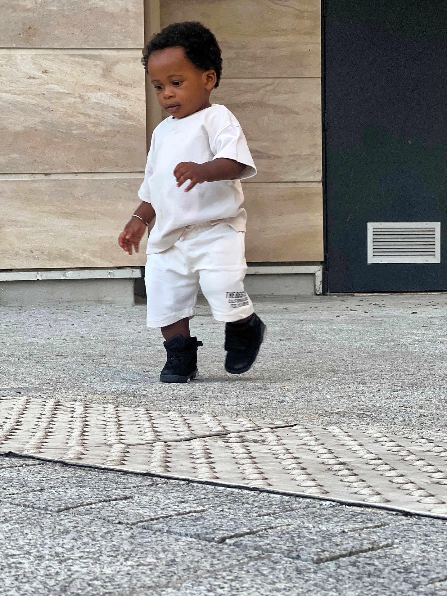Nathan participe au concours pour gagner de l'argent avec cette photo : toddler, child, walking, white_clothing, black_shoes, outdoor, concrete, urban, door, wall, vent, shorts, tshirt, person, young_child, curly_hair, side_view, casual, daylight, focused
