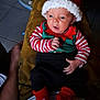 baby, infant, child, christmas_hat, red_hat, striped_shirt, festive_clothing, candy_cane_slippers, black_pants, person, sitting, indoor, legs, feet, table, books, floor_tiles, hand, curious_expression, holiday