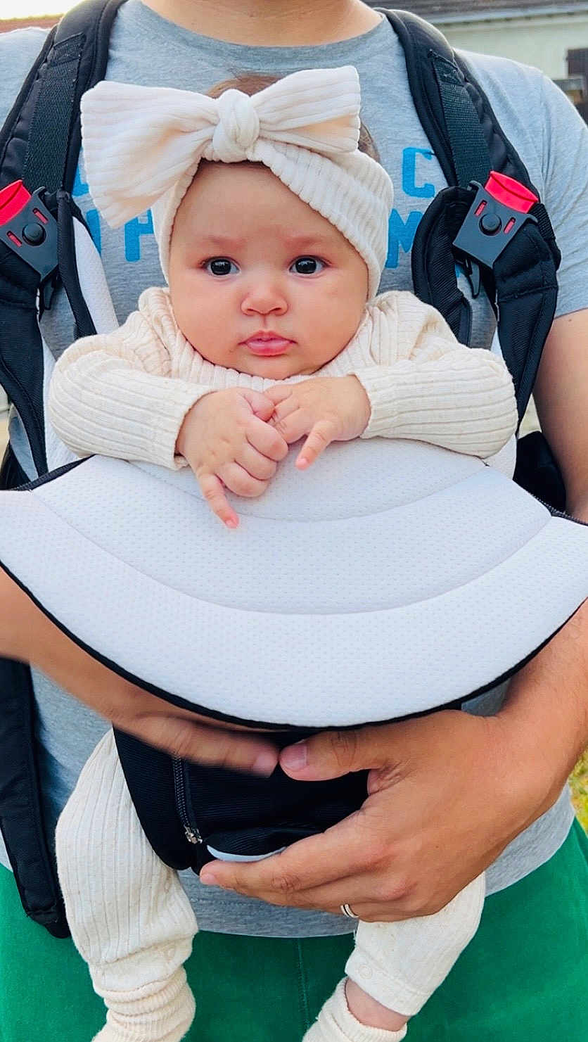 Melissa Léa participe au concours pour gagner de l'argent avec cette photo : baby, child, infant, baby_carrier, headband, bow, hands, clothing, adult, person, outdoor, green_pants, gray_shirt, holding, cute, face, expression, closeup, portrait, human