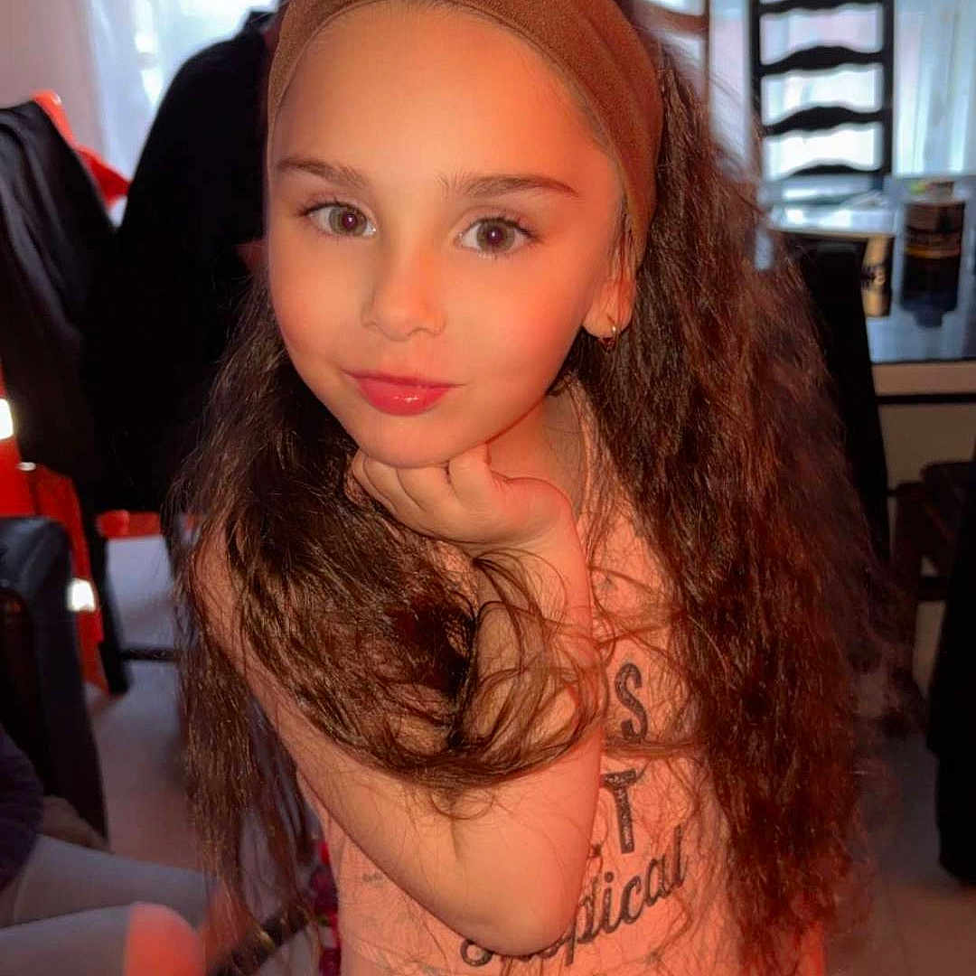 Elvina participe au concours pour gagner de l'argent avec cette photo : casual_clothing, chair, child, curly_hair, curtain, cute, face, girl, hand_on_chin, headband, home, indoor, long_hair, person, portrait, posing, smiling, table, window, young