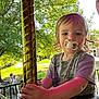 child, toddler, pacifier, carousel, pole, outdoor, park, sunlight, greenery, person, photography, summer, playground, fun, holding, seatbelt, shorts, tshirt, face, hair