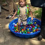 child, toddler, ball_pool, colorful_balls, outdoor, sunlight, clothing, mickey_mouse, life_vest, curly_hair, person, footwear, grill, concrete, grass, adult, pants, shoes, play, summer