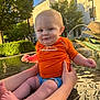 baby, child, infant, orange_shirt, fountain, water, hands, outdoor, sunlight, greenery, building, smile, legs, feet, playful, summer, daylight, person, support, water_spray