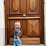 toddler, child, smiling, standing, jeans, sneakers, wooden_door, door, outdoor, happy, casual_clothing, blonde_hair, laughing, concrete, sidewalk, urban, young_child, person, candid, daylight