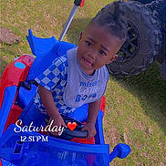 Ace is registered to the contest to win money with this photo: toddler, child, toy_car, outdoor, grass, blue, red, person, vehicle, offroad_vehicle, footwear, hairstyle, playtime, curious_expression, sunlight, daytime, casual_clothing, sitting, hand_on_steering_wheel, timestamp