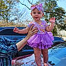 baby, child, tutu, purple, headband, car, hood, adult_hand, outdoor, trees, sky, smiling, standing, clothing, person, cute, infant, daylight, support, nature