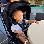Gaby participe au concours pour gagner de l'argent avec cette photo : arm, baby, black_stroller, blue_eyes, blurred_background, cafe, casual_clothing, child, cloth, curly_hair, daylight, face, hand, outdoor, pacifier, person, seatbelt, stroller, sunlight, table