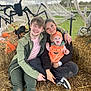 baby, child, young_adult, halloween, pumpkin_costume, spider_decorations, hay_bales, outdoor, smiling, family, festive, costume, casual_clothing, web_decor, grass, fence, people, sitting, happy, portrait