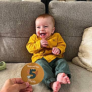 Mahé a rejoint le concours — aidez-le/la à gagner de superbes lots ! baby, child, smiling, couch, clothing, yellow_shirt, green_pants, happy, indoor, milestone_card, hand, feet, face, person, sitting, home, cozy, fabric, portrait, cute
