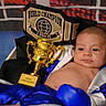 Adonis is registered to the contest to win money with this photo: baby, infant, child, boxing, boxing_gloves, championship_belt, trophy, champion, costume, blue_clothing, white_clothing, portrait, sitting, indoor, sports, celebration, gold, black_background, serious_expression, photography