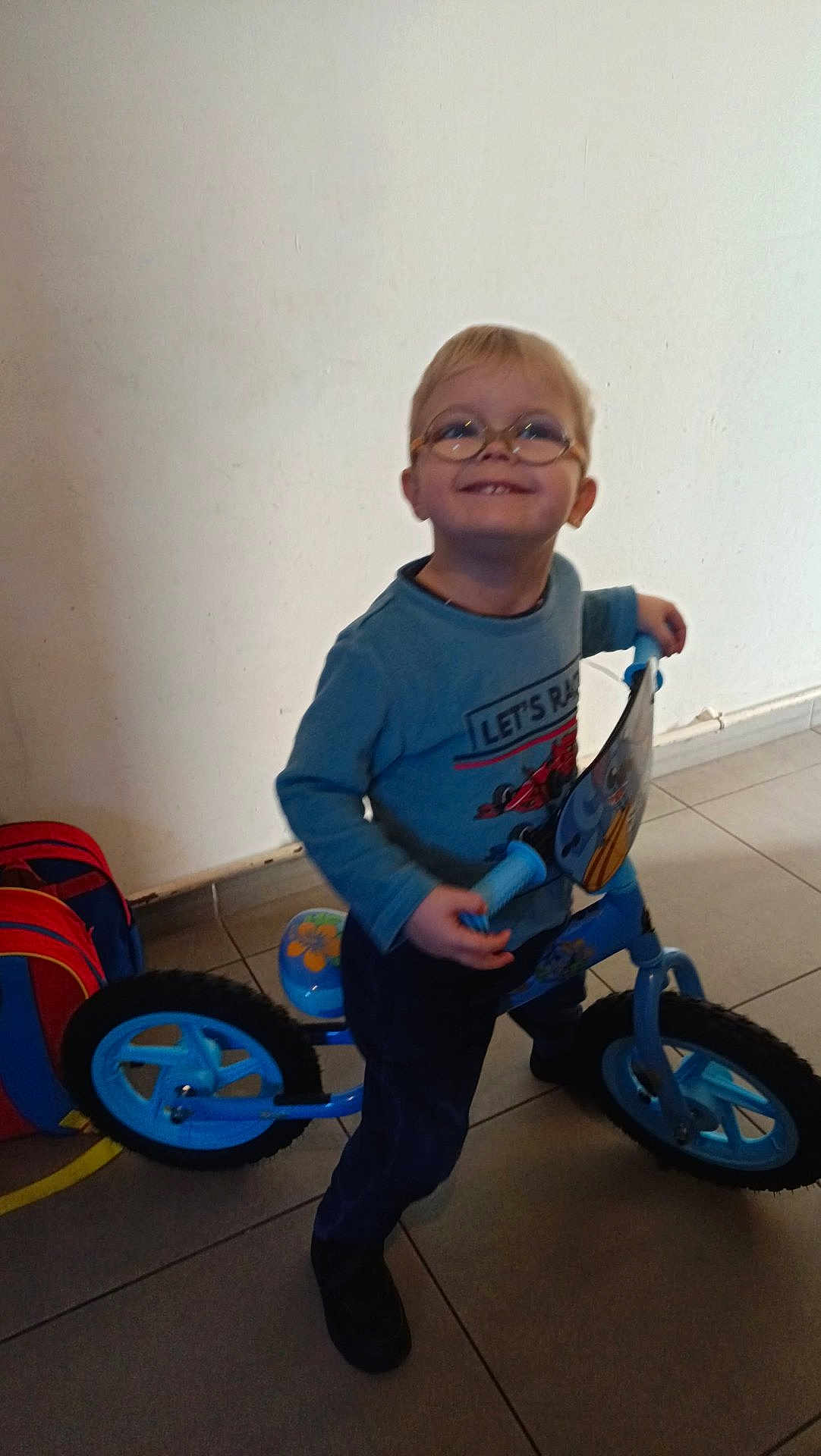 Sylvain Junior participe au concours pour gagner de l'argent avec cette photo : backpack, balance_bike, blue_bike, casual_clothing, child, floor_tiles, footwear, glasses, happy, indoor, pants, person, play, portrait, smiling, standing, sweater, toddler, toy, young_child