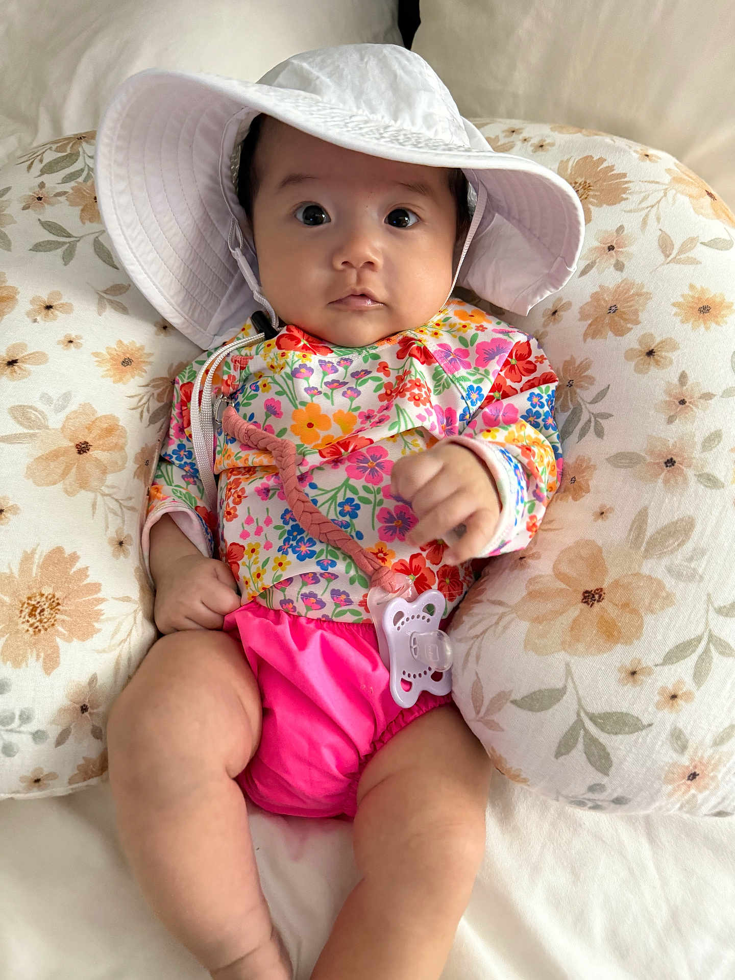 Viveeana is registered to the contest to win money with this photo: baby, infant, hat, floral, colorful_clothing, pacifier, pillow, cute, indoors, soft_fabric, child, pink, white, skin, person, sitting, young, closeup, face, relaxing