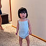 toddler, child, indoor, carpet, barefoot, onesie, wall, wooden_baseboard, person, short_hair, standing, neutral_expression, home_interior, adult_partial, floor, light_blue_clothing, young_child, asian_child, curious, small_person