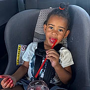 Amara is registered to the contest to win money with this photo: child, car_seat, drink, straw, red_drink, toddler, hair_tie, white_shirt, sitting, car_interior, seatbelt, warning_label, happy, smiling, fingers, shorts, black_hair, eating, portrait, young