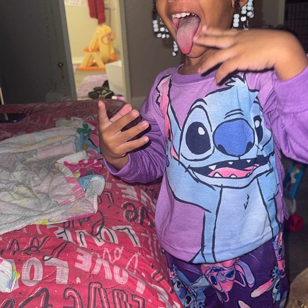 Amara is registered to the contest to win money with this photo: beads, bed, braids, child, clothing, doorway, floor, hand_gesture, happy, indoor, love_text, pajamas, person, quilt, room, smile, stitch, tongue_out, towel, toy