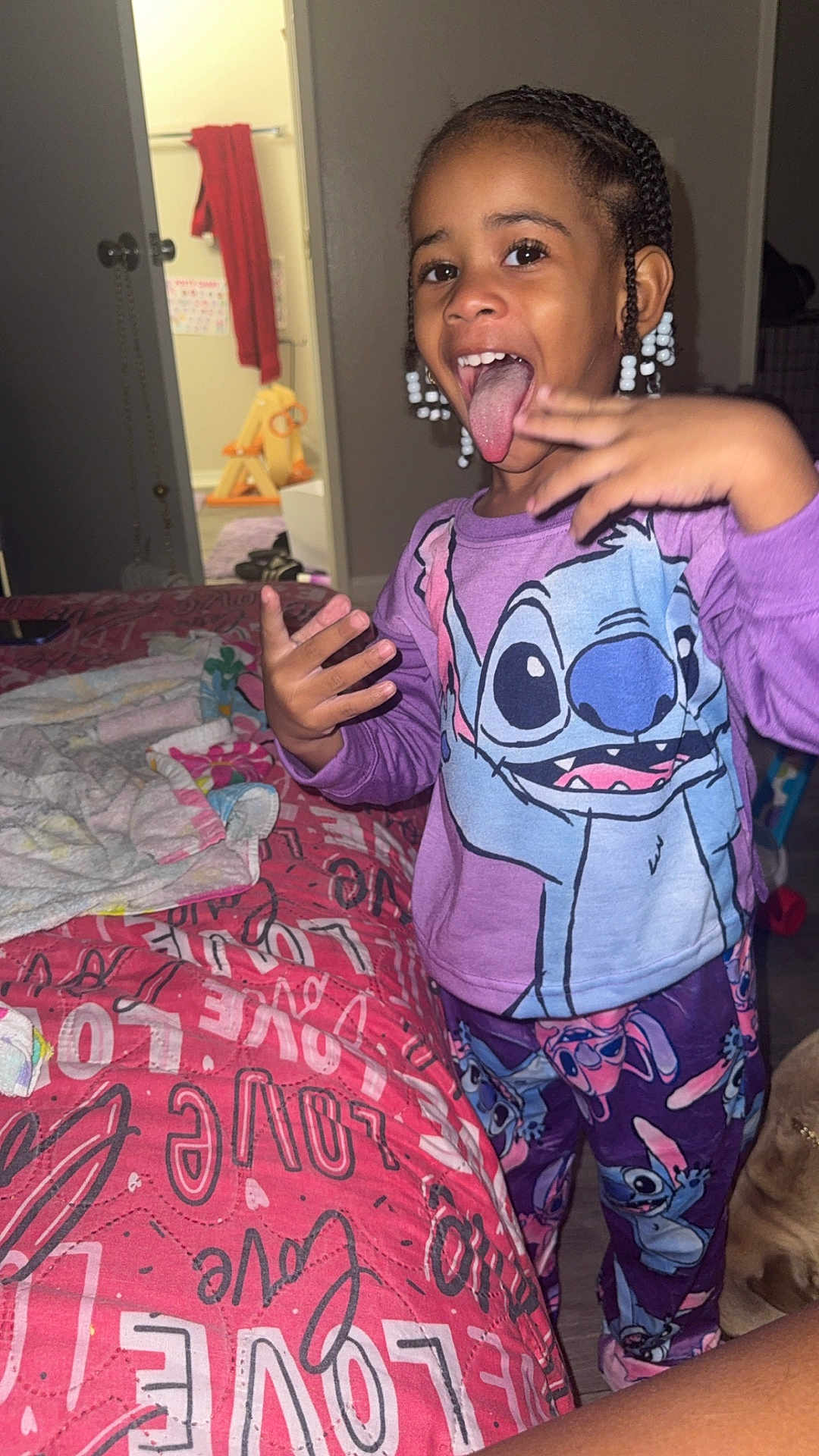 Amara is registered to the contest to win money with this photo: child, pajamas, stitch, tongue_out, bed, quilt, love_text, hand_gesture, smile, indoor, room, doorway, towel, toy, braids, beads, floor, happy, person, clothing