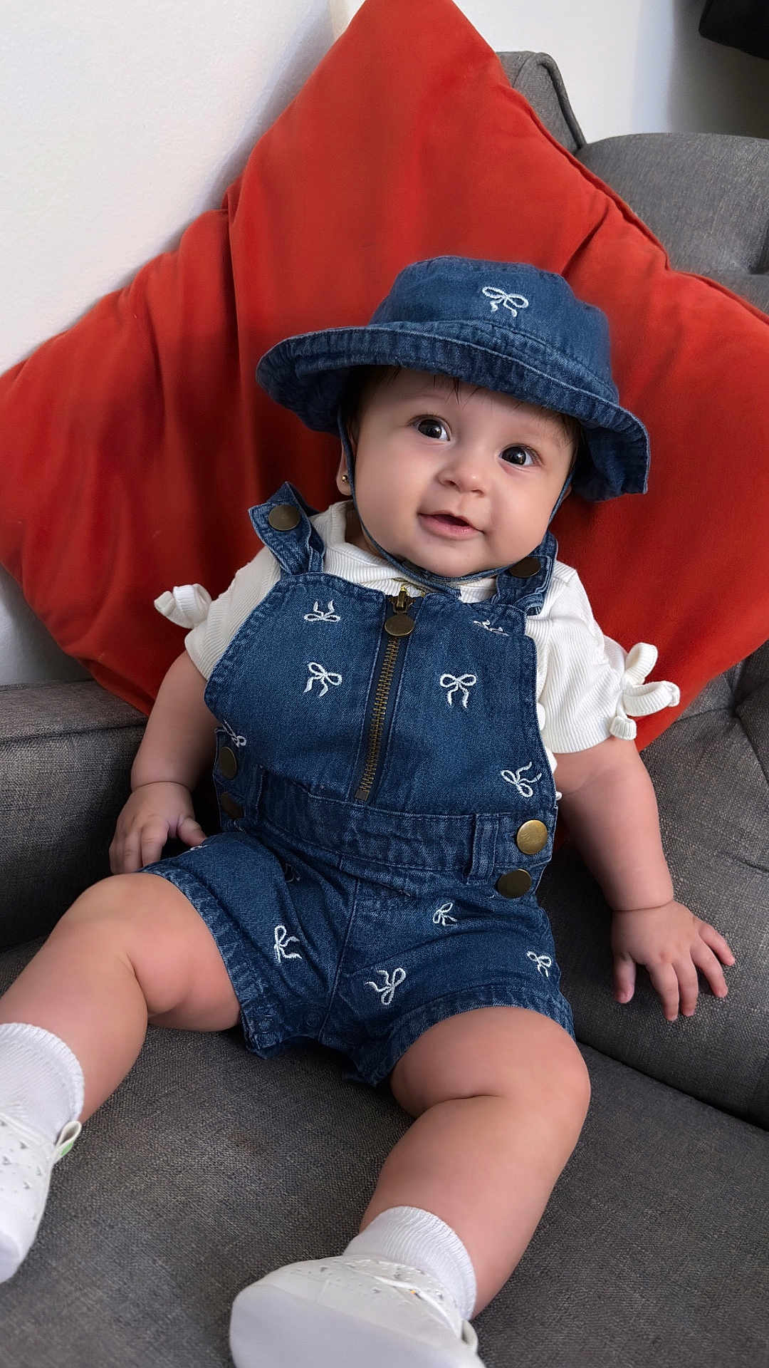 Aysha is registered to the contest to win money with this photo: baby, denim, overalls, hat, smile, couch, orange_pillow, white_shoes, white_socks, indoor, child, cute, portrait, happy, seated, clothing, face, person, infant, home