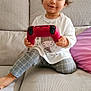 casual_clothing, child, controller, couch, cute, game_controller, grey_pants, happy, indoor, living_room, person, pillow, playful, ponytail, purple_pillow, seated, smiling, toddler, white_shirt, young_child