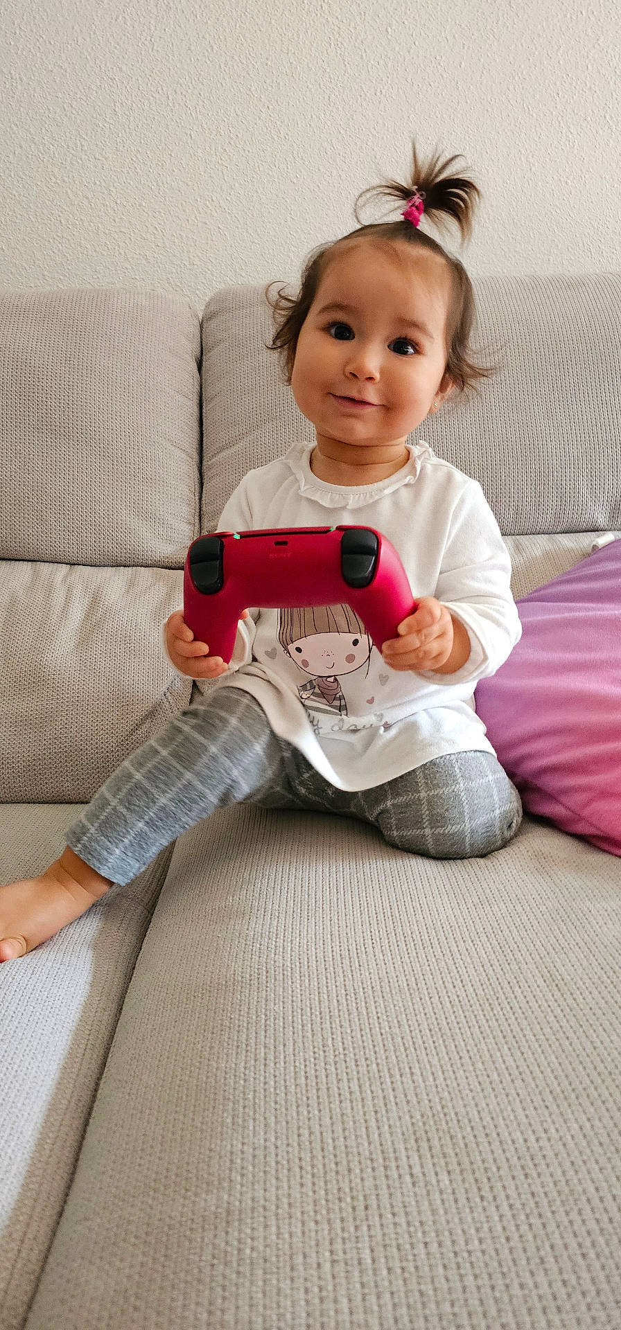 Emma a rejoint le concours — aidez-le/la à gagner de superbes lots ! casual_clothing, child, controller, couch, cute, game_controller, grey_pants, happy, indoor, living_room, person, pillow, playful, ponytail, purple_pillow, seated, smiling, toddler, white_shirt, young_child
