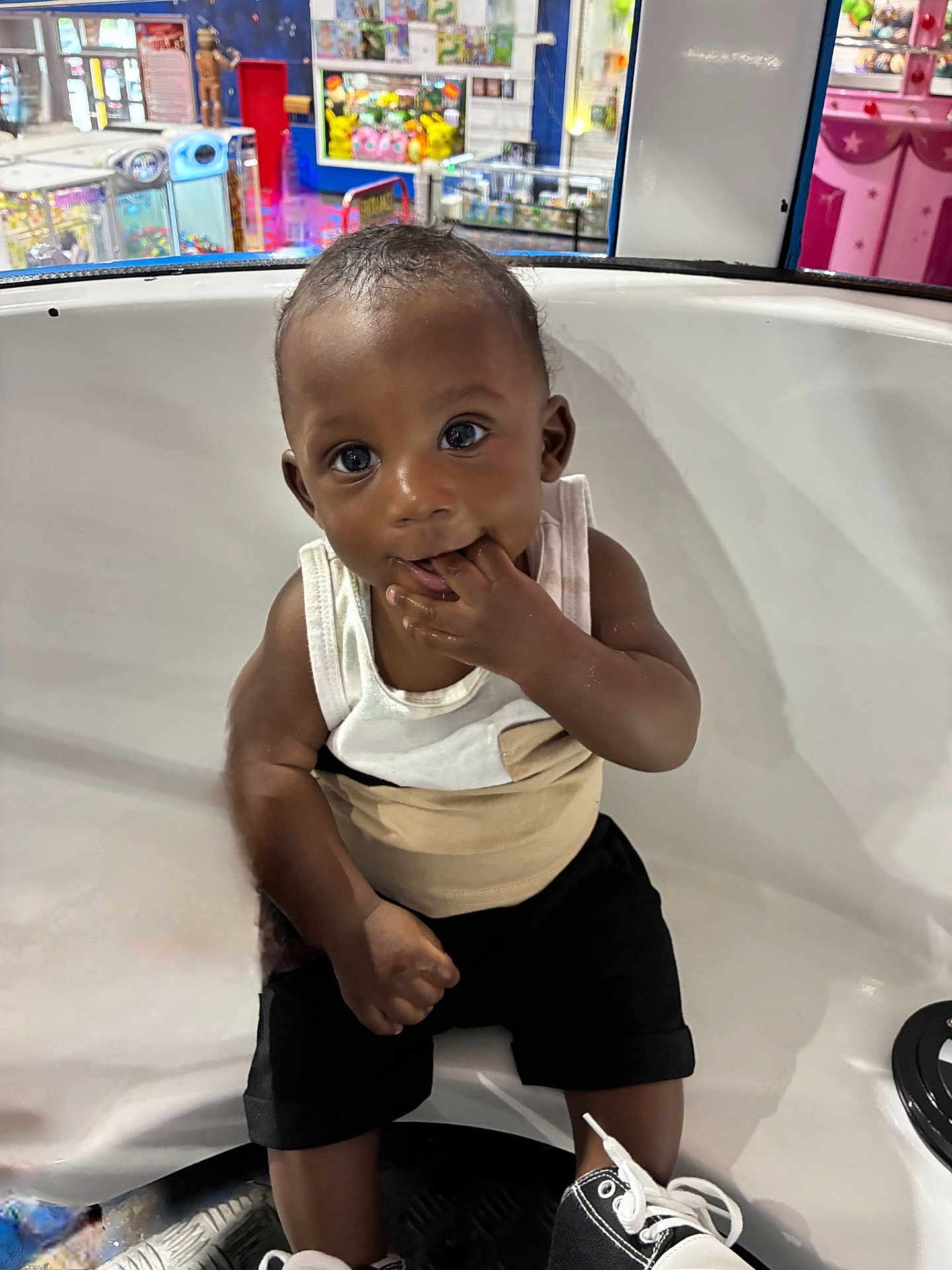Jaylen joined the competition — help win amazing prizes! baby, toddler, child, sitting, playing, hand_in_mouth, white_tank_top, black_shorts, sneakers, arcade, colorful_background, indoor, play_area, smiling, cute, curious, face, person, seat, fun