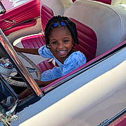Rhyli joined the competition — help win amazing prizes! blue_dress, braids, car_interior, child, classic_car, girl, hair_clips, happy, outdoor, person, pink, portrait, seat, smile, steering_wheel, sunlight, vehicle, vintage_car, white, window