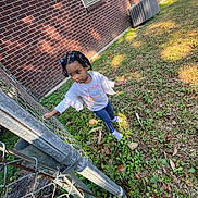 Nevaeh joined the competition — help win amazing prizes! child, outdoor, grass, fence, brick_wall, sunlight, hair_clips, curious, blue_clothing, long_sleeve_shirt, pants, socks, person, nature, exploring, shadows, daylight, young_child, standing, playful