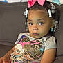 Jenasis is registered to the contest to win money with this photo: child, girl, braids, beads, pink_bow, tshirt, cartoon_print, couch, indoor, portrait, young_child, sitting, holding_object, curious_expression, casual_clothing, african_american, home, relaxed, cute, young