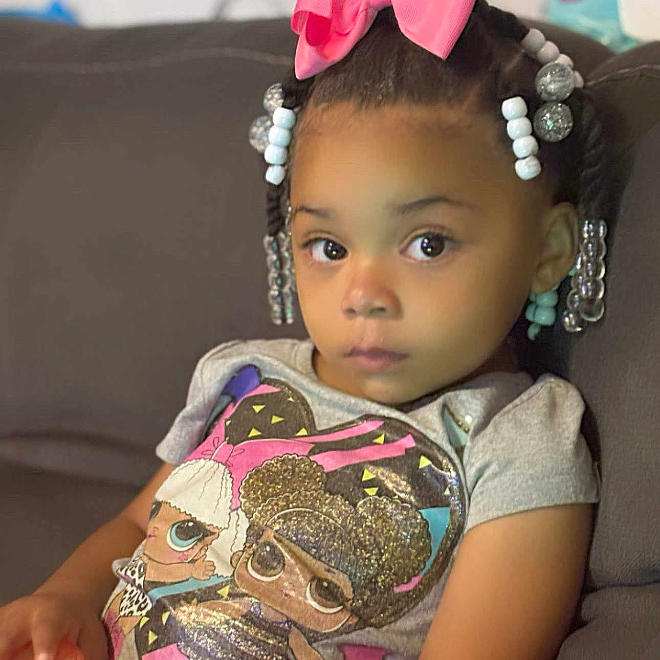 Jenasis is registered to the contest to win money with this photo: african_american, beads, braids, cartoon_print, casual_clothing, child, couch, curious_expression, cute, girl, holding_object, home, indoor, pink_bow, portrait, relaxed, sitting, tshirt, young, young_child