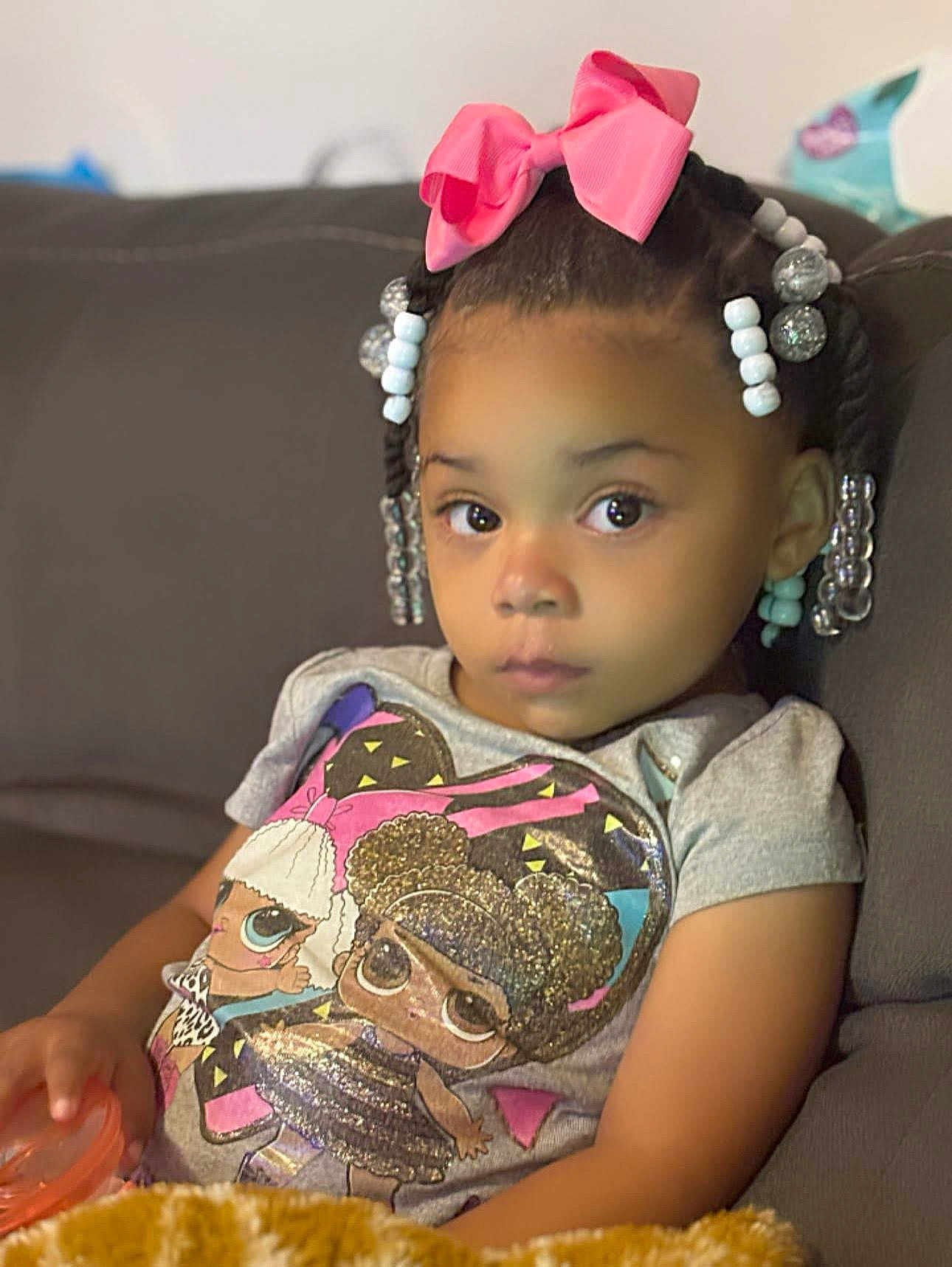 Jenasis is registered to the contest to win money with this photo: child, girl, braids, beads, pink_bow, tshirt, cartoon_print, couch, indoor, portrait, young_child, sitting, holding_object, curious_expression, casual_clothing, african_american, home, relaxed, cute, young