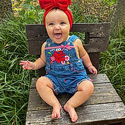 Landrie is registered to the contest to win money with this photo: baby, child, red_bow, denim_overalls, smiling, tongue_out, wooden_chair, outdoor, greenery, grass, happy, cute, infant, nature, portrait, sitting, headband, playful, person, summer