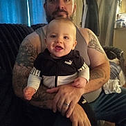Bradley is registered to the contest to win money with this photo: baby, man, tattoo, tank_top, puma_jacket, smile, couch, living_room, curtains, indoor, person, child, holding, casual_clothing, beard, seated, home, furniture, happy, portrait