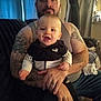 baby, man, tattoo, tank_top, puma_jacket, smile, couch, living_room, curtains, indoor, person, child, holding, casual_clothing, beard, seated, home, furniture, happy, portrait