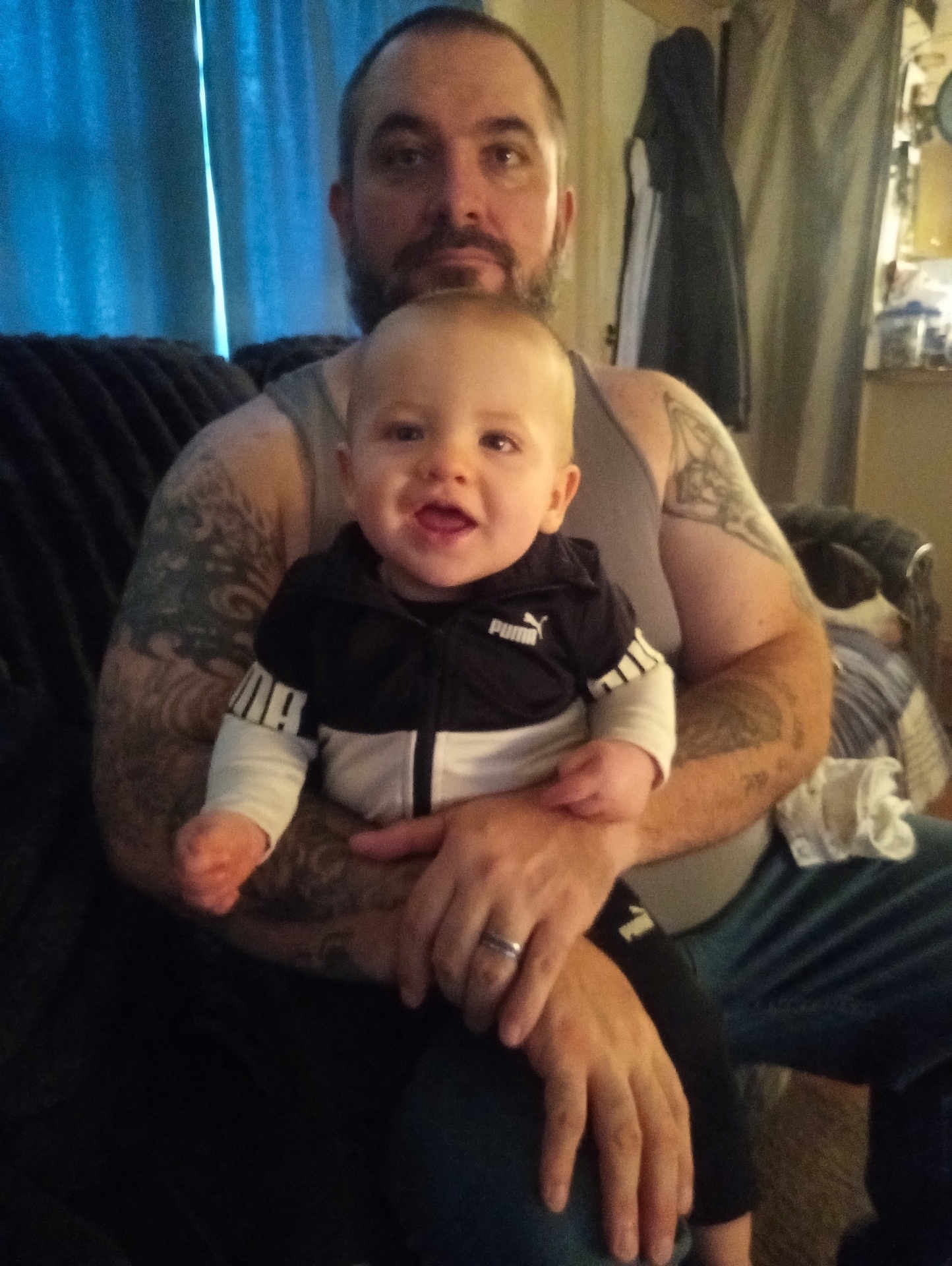 Bradley is registered to the contest to win money with this photo: baby, man, tattoo, tank_top, puma_jacket, smile, couch, living_room, curtains, indoor, person, child, holding, casual_clothing, beard, seated, home, furniture, happy, portrait