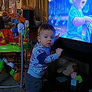 Bradley is registered to the contest to win money with this photo: child, toddler, standing, baby_on_tv, television, toys, stuffed_animal, play_center, blocks, carpet, curtains, sweatshirt, pants, living_room, wall_art, posters, furniture, play_toy, screen, indoor
