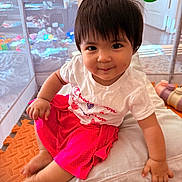 Cyra is registered to the contest to win money with this photo: toddler, child, baby, smiling, sitting, playpen, toy, cushion, clothing, indoors, cute, happy, person, face, short_hair, pink_skirt, white_tshirt, floor_mat, play_area, young_child
