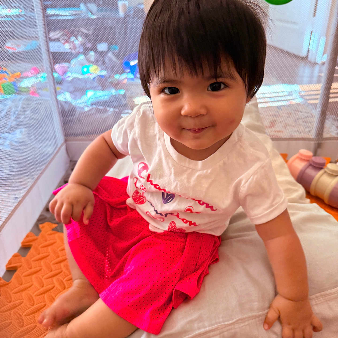 Cyra is registered to the contest to win money with this photo: toddler, child, baby, smiling, sitting, playpen, toy, cushion, clothing, indoors, cute, happy, person, face, short_hair, pink_skirt, white_tshirt, floor_mat, play_area, young_child