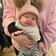 Mazie joined the competition — help win amazing prizes! baby, child, person, pink_sweatshirt, knit_hat, pom_poms, smiling, bib, striped_pants, hand, indoor, floor_tiles, holding, cute, warm_clothing, black_shirt, happy, portrait, infant, cozy