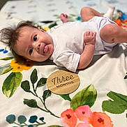 Gracelyn- Claire joined the competition — help win amazing prizes! baby, infant, tongue_out, white_clothing, floral_blanket, colorful, cute, lying_down, three_months, wooden_sign, face, skin, hand, foot, expression, child, indoors, soft_light, portrait, playful