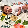 baby, infant, tongue_out, white_clothing, floral_blanket, colorful, cute, lying_down, three_months, wooden_sign, face, skin, hand, foot, expression, child, indoors, soft_light, portrait, playful