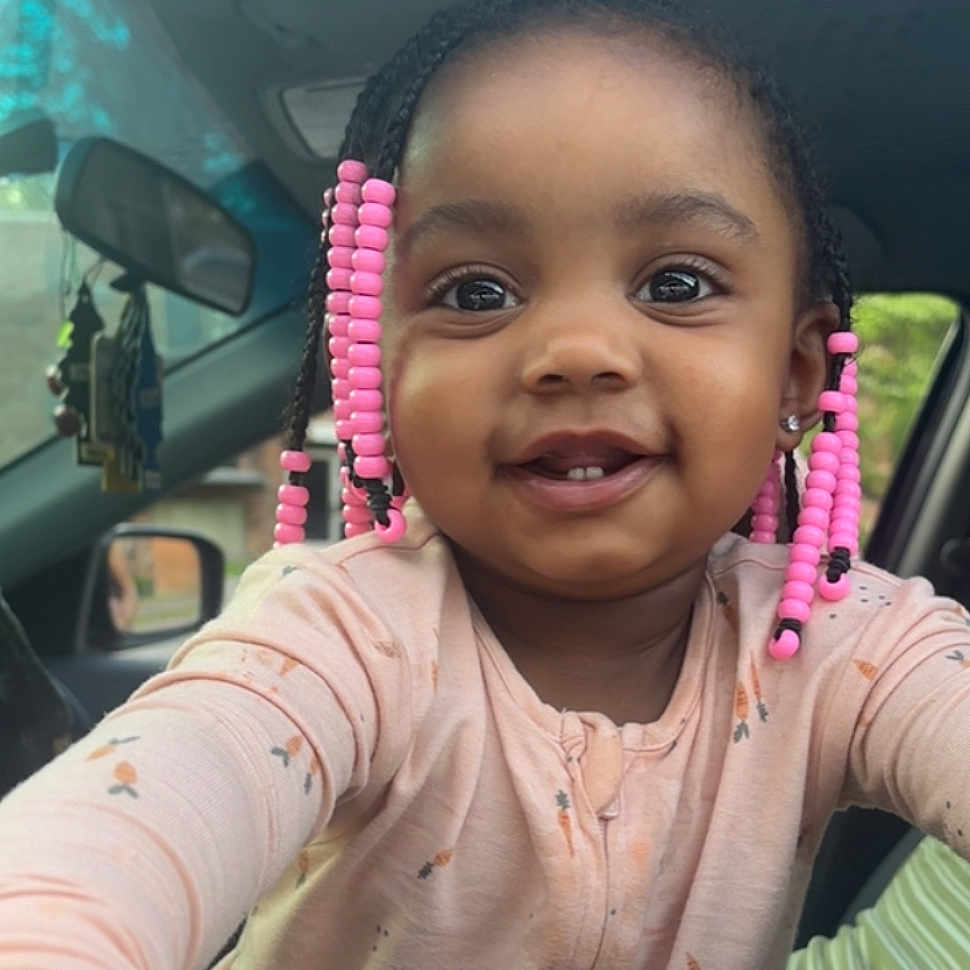 Barbie is registered to the contest to win money with this photo: braids, car_interior, child, clothing, cute, door, eyes, face, happy, indoors, long_sleeves, mirror, person, pink_beads, portrait, seat, smiling, toddler, window, young_child